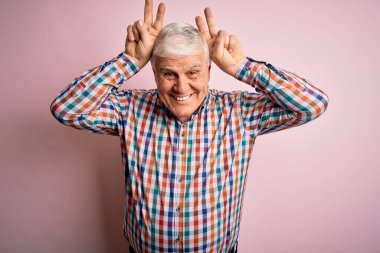 Üst düzey yakışıklı, parlak gömlekli, pembe arka planda, komik ve çılgın pozlar veren, parmakları tavşan kulaklı, neşeli gülümseyen bir adam.