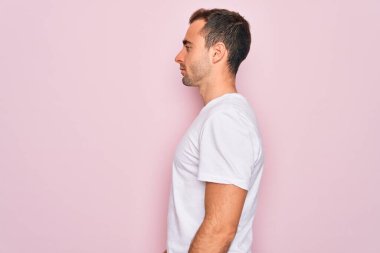 Yakışıklı, mavi gözlü, günlük beyaz tişört giyen, pembe arka planda duran, yan tarafa bakan, rahat pozu veren, kendine güvenen, gülümseyen bir adam..