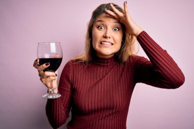 Genç, güzel, sarışın bir kadın izole edilmiş pembe arka planda bir bardak kırmızı şarap içiyor. Elleri başının üstünde, utanç ve şaşkınlık dolu, sinirli ve hayal kırıklığına uğramış bir halde. Hata için korku ve üzüntü.
