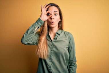 Sarı arka planda yeşil tişört giyen, mavi gözlü genç, güzel sarışın kadın şaşırmış yüz ifadesiyle şaşırmış, parmaklarından bakan bir jest yapıyor. İnançsız ifade.