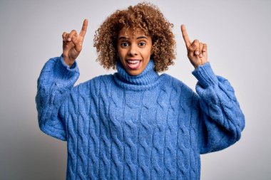 Beyaz arka planda balıkçı yaka kazak giyen güzel Afro-Amerikalı kadın. Şaşırmış, şaşırmış ve parmaklarıyla işaret ediyor ve kollarını kaldırıyordu..