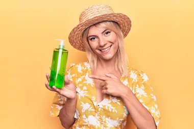 Güzel sarışın kadın tatilde. Yazlık şapka takıyor. Aloe vera kremalı bir şişe tutuyor.