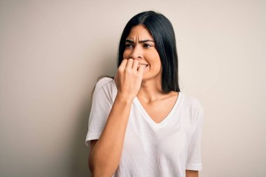 İzole edilmiş arka plan üzerinde gündelik beyaz tişört giyen genç ve güzel İspanyol kadın elleri ağız yiyen tırnaklarla stresli ve gergin görünüyor. Endişe sorunu.