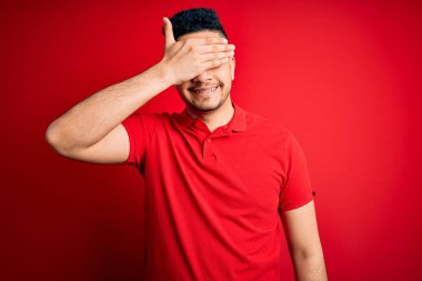 İzole edilmiş arka planda kırmızı polo giyen genç yakışıklı adam gülümseyerek ve gülerek gözleri kapayarak şaşkınlık içinde bekliyor. Kör kavram.