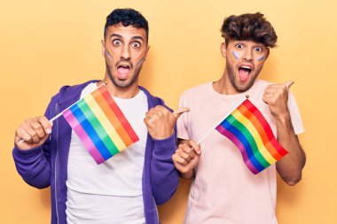 Genç eşcinsel çift ellerinde gökkuşağı lgbtq bayrakları başparmağıyla yan tarafa bakıyor mutlu bir şekilde ağzı açık gülümsüyor 