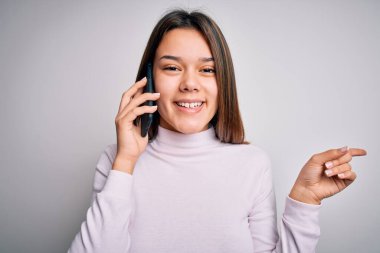 Güzel esmer kız beyaz arka planda akıllı telefondan konuşuyor elini ve parmağını yana uzatarak mutlu bir şekilde işaret ediyor.