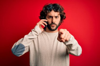 Sakallı yakışıklı bir adam kırmızı arka planda akıllı telefondan konuşuyor. Kamerayı işaret ediyor ve sen, el işareti, ön taraftan olumlu ve kendinden emin bir hareket.