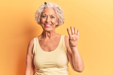 Kıdemli, gri saçlı, günlük kıyafetler giyen, dört numarayı gösteren ve gülümseyen, kendinden emin ve mutlu bir kadın.. 