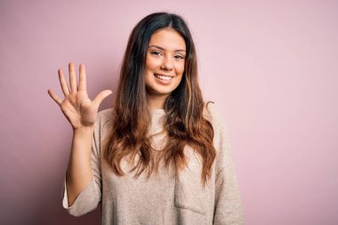 Genç, güzel esmer kadın pembe arka planda dikilip gülümseyen, kendinden emin ve mutlu gülümseyen beş numarayı işaret ediyor..