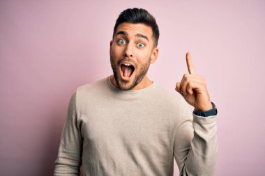 İzole edilmiş pembe arka plan üzerinde duran sıradan bir süveter giyen yakışıklı genç adam başarılı bir fikirle işaret ediyor. Heyecanlı ve mutlu. Bir numara..