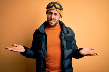 Sakallı, kar maskesi takan, kar gözlüklü, kolları açık ve kafası karışık genç, yakışıklı bir kayakçı..