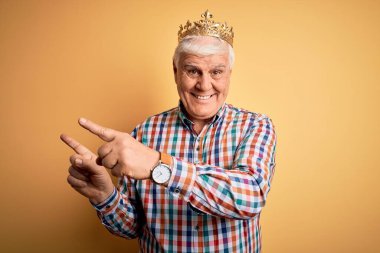 Sarı arka planda kralın altın tacını takmış yaşlı yakışıklı adam gülümsüyor ve iki eliyle ve parmaklarıyla kameraya bakıyor..