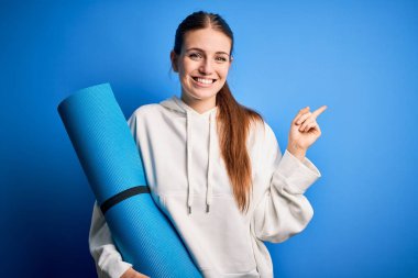 Genç, güzel, kızıl saçlı, sportif bir kadın mavi arka planda yoga minderi tutuyor.