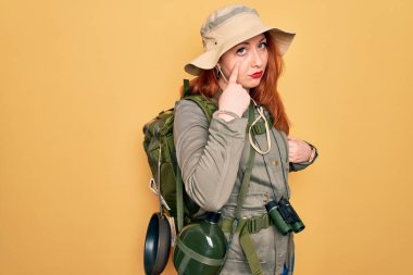 Sırt çantası ve sarı arka planda şapka takan kızıl saçlı genç bir kadın. Gözünü dikip jestini izliyor. Şüpheli bir ifade.