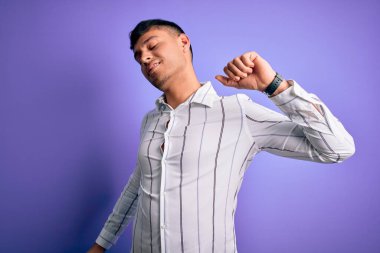 Zarif iş gömleği giyen genç yakışıklı İspanyol adam mor arka planda duruyor, geriniyor, yorgun ve rahat, uykulu ve sabahın erken saatlerinde esniyor.