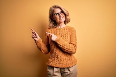 Orta yaşlı güzel sarışın kadın sarı arka planda rahat süveter ve gözlük takıyor gülümsüyor ve kameraya bakıyor iki eliyle ve parmaklarıyla yandan işaret ediyor..