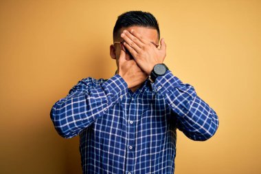 Sarı arka plan üzerine gündelik gömlek ve gözlük takan genç, yakışıklı Latin adam gözleri ve ağzı kapalı, şaşırmış ve şok olmuş. Duygularını gizliyor.