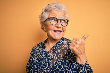 Sarı arka planda günlük gömlek ve gözlük takan yaşlı, güzel, gri saçlı kadın gülümsüyor ve baş parmakla yüzünü gösteriyor..