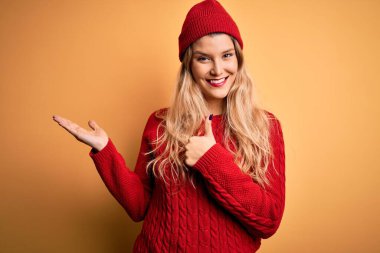 Genç, sarışın, günlük süveter giyen, beyaz arka planda yün şapka takan, avuçlarını gösteren, baş parmaklarıyla hareket eden, mutlu ve neşeli gülümseyen bir kadın.