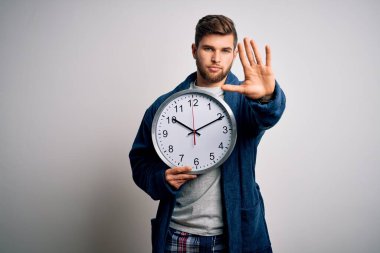 Sakallı ve mavi gözlü genç bir adam pijama giyiyor. Elinde kocaman bir saat var. Elinde dur işareti var.