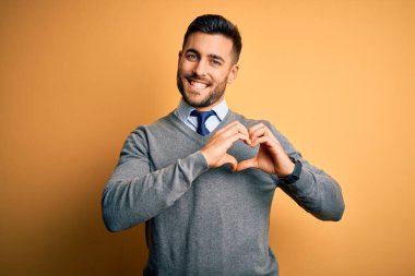 Zarif bir süveter ve sarı arka planda kravat takan genç yakışıklı iş adamı. Aşkla gülümsüyor. Elleriyle kalp sembolü ve şekli gösteriyor. Romantik konsept.