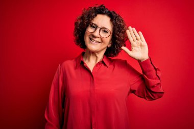 Orta yaşlı güzel kıvırcık saçlı kadın kırmızı arka planda günlük gömlek ve gözlük takıyor. Mutlu ve güler yüzlü, dostça karşılama jesti yapıyor.