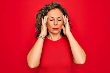 Orta yaşlı, esmer, sıradan tişörtler giyen bir kadın. Kırmızı arka planda duruyor. Başı ağrıyor çünkü stres altında. Migren ağrısı..