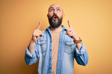 Sıradan kot ceket ve çizgili tişört giyen sakallı yakışıklı kel adam şaşkın şaşkın yukarı bakıyor ve parmaklarıyla işaret ediyor ve kollarını kaldırıyor..