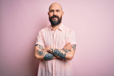 Sakallı ve dövmeli yakışıklı, pembe arka planda rahat bir gömlek giyen, kollarını kavuşturup kameraya bakan, gülümseyen, kel bir adam. Pozitif kişi.