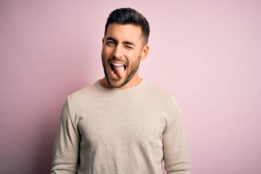 İzole edilmiş pembe arka planda dikilen sıradan süveter giyen genç yakışıklı adam komik bir ifadeyle dilini dışarı çıkartıyor. Duygu kavramı.