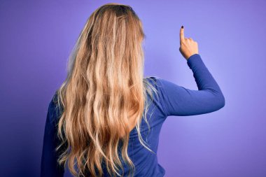 İzole edilmiş mor arka plan üzerine gündelik tişört giyen genç ve güzel sarışın bir kadın parmak eliyle öne doğru poz veriyor.