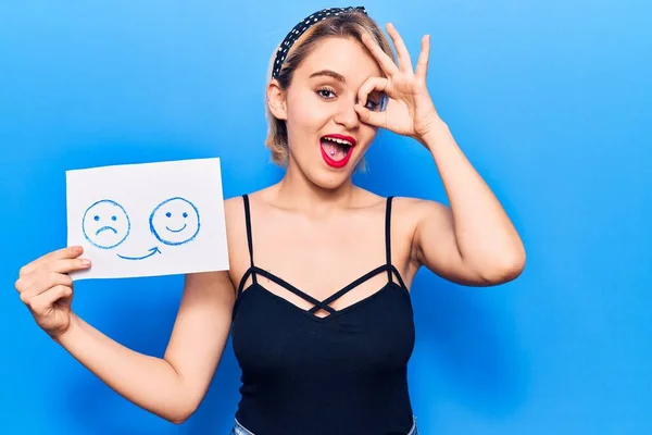 Young beautiful blonde woman holding sad to happy emotion paper smiling happy doing ok sign with hand on eye looking through fingers