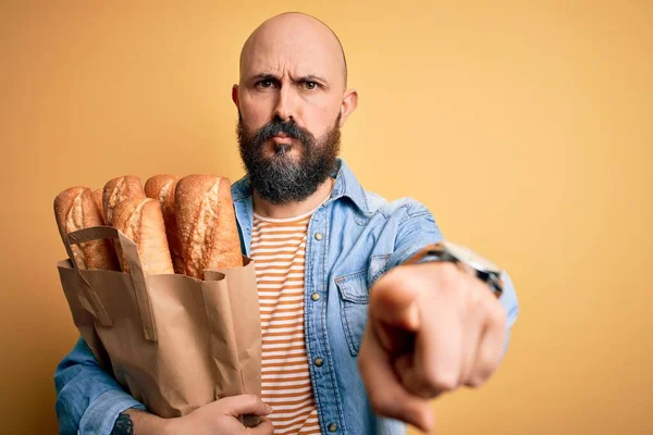 Handsome bald man with beard holding paper bag with bread over yellow background pointing with finger to the camera and to you, hand sign, positive and confident gesture from the front