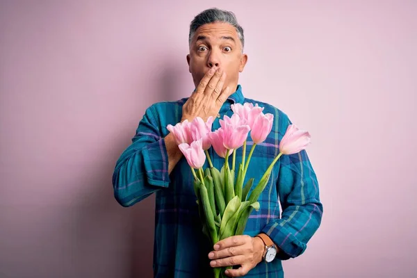 Middle age grey-haired man holding natural bouquet of pink tulips over isolated background cover mouth with hand shocked with shame for mistake, expression of fear, scared in silence, secret concept