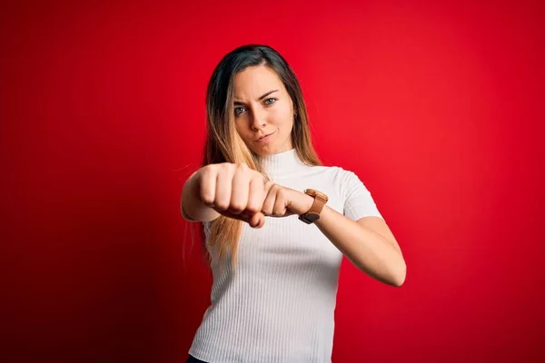 Beautiful blonde woman with blue eyes wearing casual white t-shirt over red background Punching fist to fight, aggressive and angry attack, threat and violence