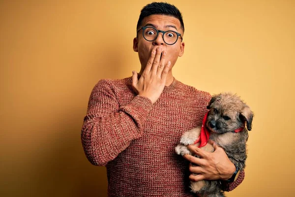 Young handsome latin man holding cute puppy pet over isolated yellow background cover mouth with hand shocked with shame for mistake, expression of fear, scared in silence, secret concept