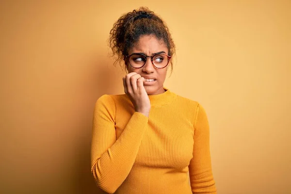 黄色の背景にセーターと眼鏡を身に着けている若い美しいアフリカ系アメリカ人の少女は 口の爪をかむ上で手で強調し 緊張している 不安の問題 — ストック写真