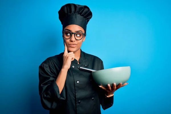 Young african american chef girl wearing cooker uniform and hat using whisk and bowl serious face thinking about question, very confused idea