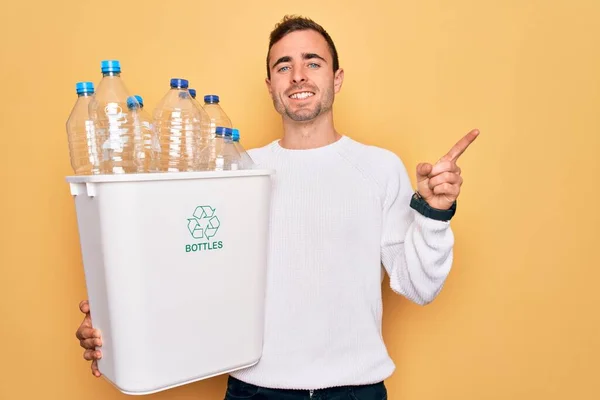 青い目の若いですハンサムな男リサイクル保持Wastebasketとともにプラスチックボトル笑顔幸せなポインティングで手と指で側面 — ストック写真