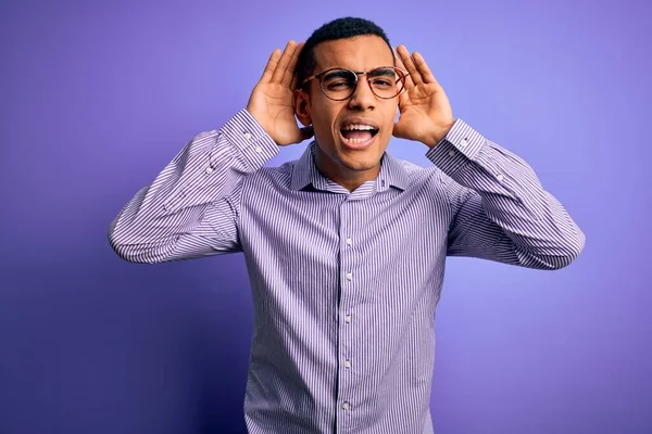 英俊的非洲裔美国男人 穿着条纹衬衫 戴着紫色背景的眼镜试图听到两只手对著耳朵的手势 好奇的流言 听力问题 — 图库照片