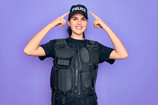 年轻的女警察身穿防弹背心制服 穿着紫色背景的制服 微笑着用双手指指着头 好主意或好主意 好记性 — 图库照片