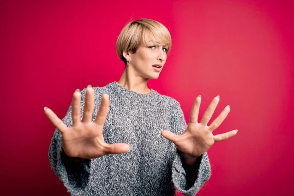 Young blonde woman with modern short hair wearing casual sweater over pink background doing stop gesture with hands palms, angry and frustration expression
