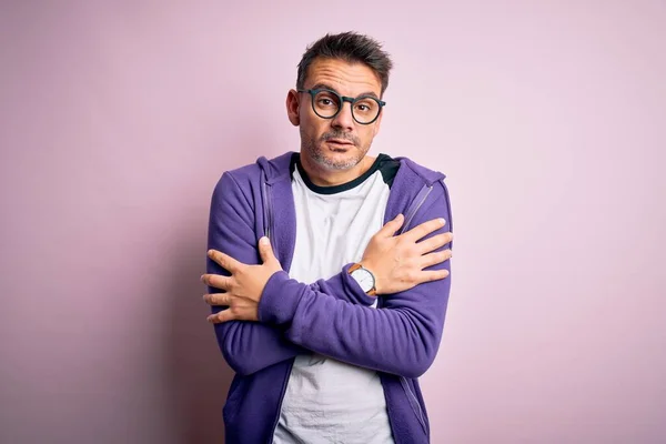 Young handsome man wearing purple sweatshirt and glasses standing over pink background shaking and freezing for winter cold with sad and shock expression on face