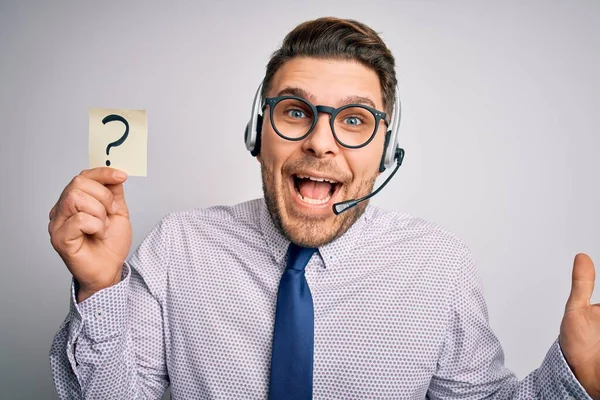 Young call center operator business man with blue eyes holding paper note with question mark very happy and excited, winner expression celebrating victory screaming with big smile and raised hands