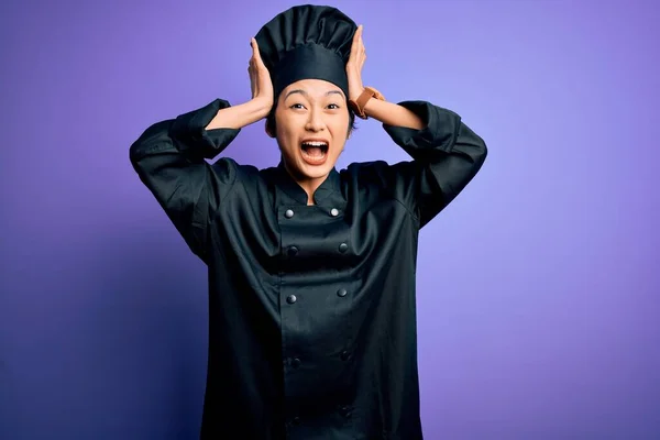 若いです美しいです中国人シェフ女性身に着けています炊飯器の制服と帽子以上紫の背景クレイジーと恐怖で手で頭 恐怖と驚きのショックでオープン口 — ストック写真