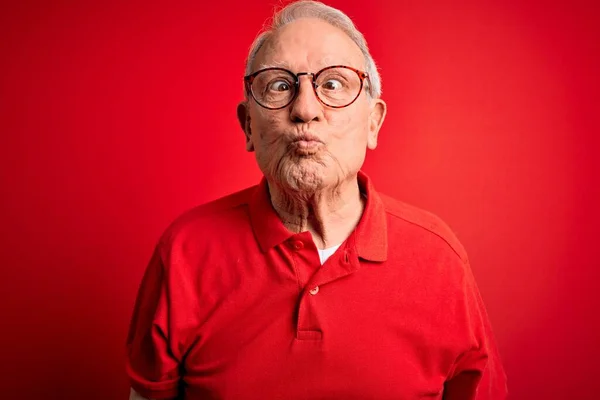 Grey haired senior man wearing glasses and casual t-shirt over red background making fish face with lips, crazy and comical gesture. Funny expression.