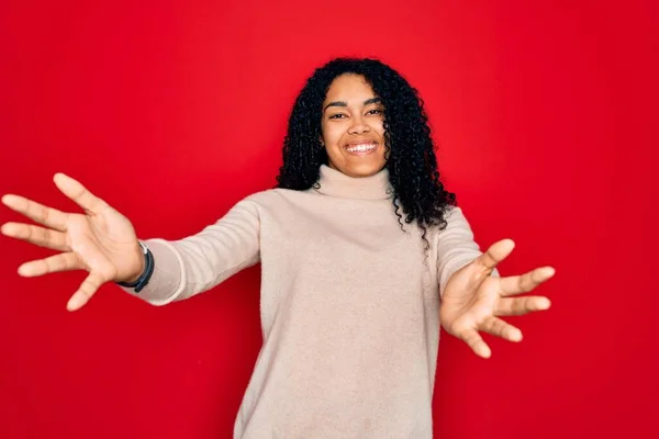 Young african american curly woman wearing casual turtleneck sweater over red background looking at the camera smiling with open arms for hug. Cheerful expression embracing happiness.