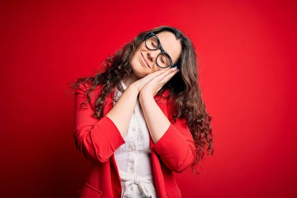 Young beautiful woman with curly hair wearing jacket and glasses over red background sleeping tired dreaming and posing with hands together while smiling with closed eyes.
