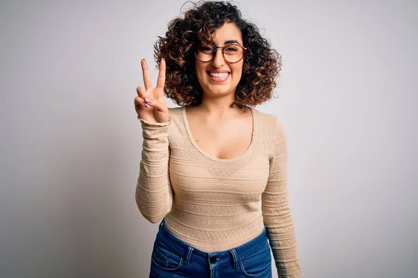 白い背景にカジュアルなTシャツと眼鏡を身に着けている若い美しい巻きアラブの女性は 勝利のサインを行う指を示すカメラを見て微笑んだ 第二番 — ストック写真