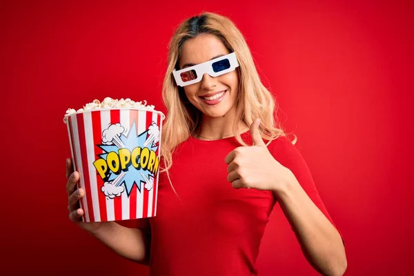 Young beautiful blonde woman watching movie using 3d glasses eating popcorns as snack happy with big smile doing ok sign, thumb up with fingers, excellent sign